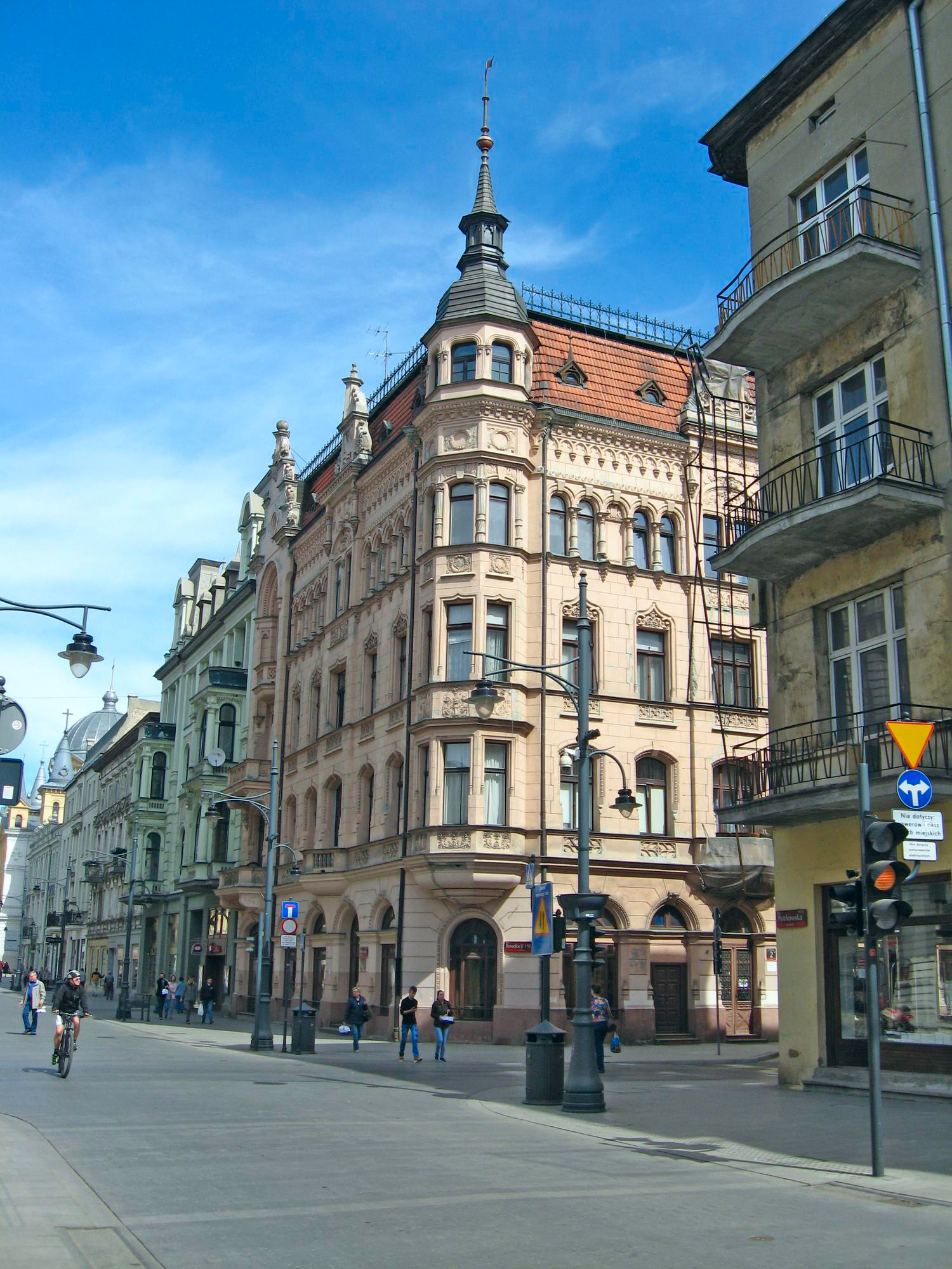 Historic European street corner with an ornate multi-story building featuring a pointed spire and red-tiled roof under a blue sky.