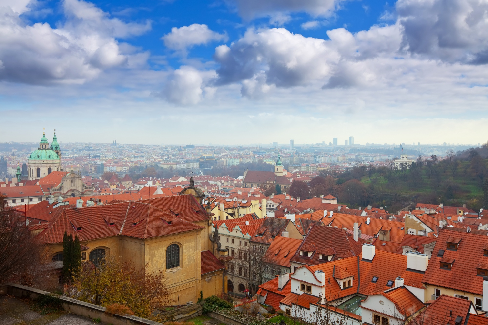 Panoramic view of a cityscape with many red-tiled roofs, historical buildings, and a large church with green domes under a cloudy blue sky.